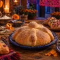 Panes de distintas formas, incluido un bollo redondo con adornos de masa, sobre una mesa con un recetario, flores y velas.